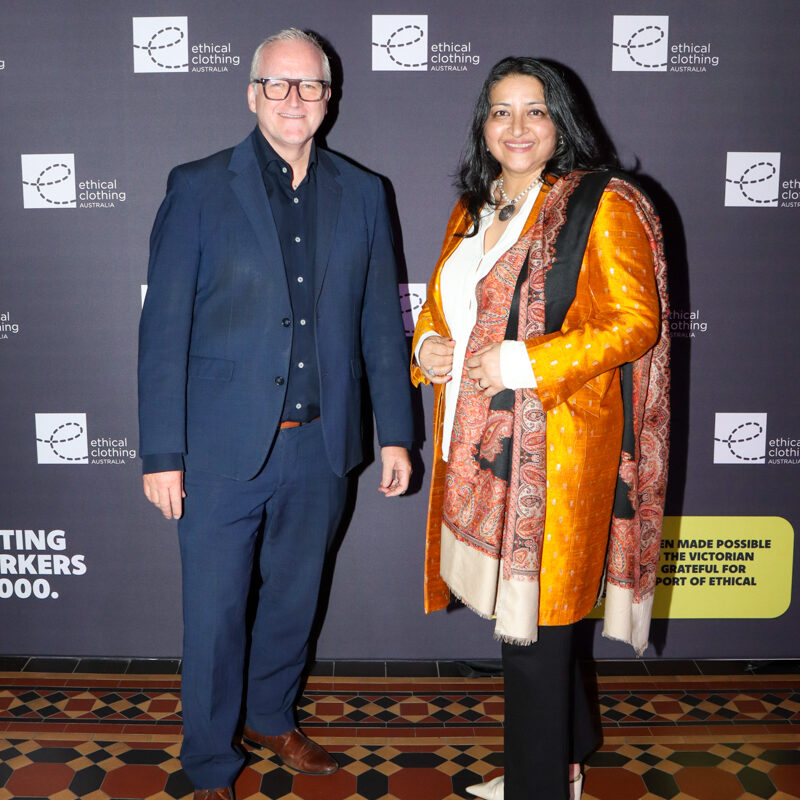 A women and a man standing in front of black media wall with a white e symbolising the Ethical Clothing Australia logo.